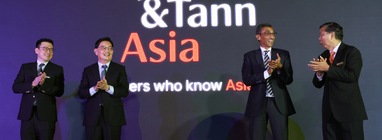 Four men in suits applaud under a '&Tann Asia' display.